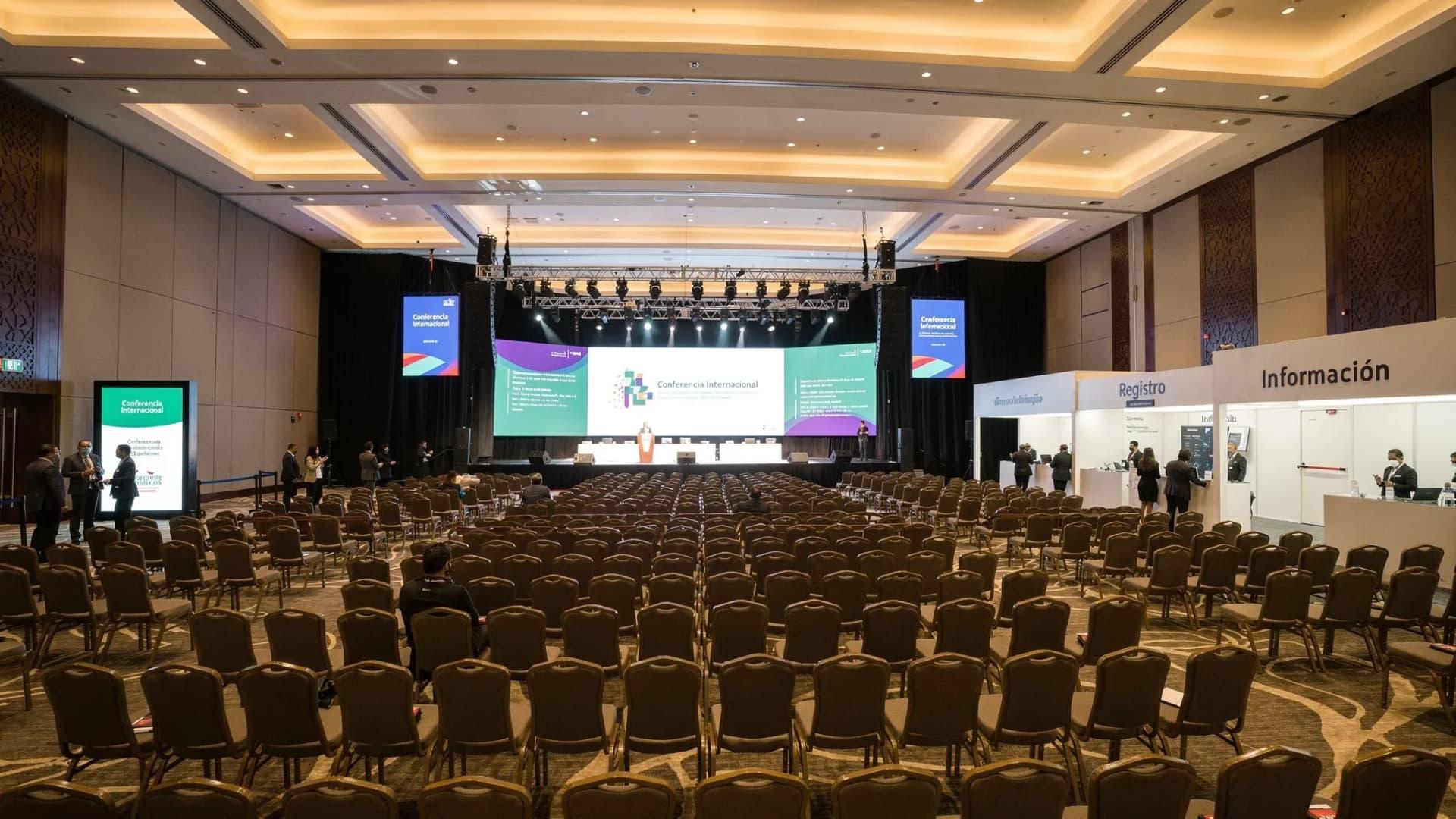 Vista interior de un centro de convenciones moderno en México, salón principal sin columnas con techo alto, montaje en teatro para gran conferencia, señalética digital, iluminación profesional y área de registro al fondo con personal de staff.